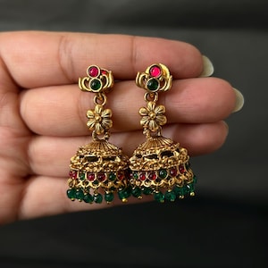 May include: A pair of gold-toned jhumka earrings featuring a bell-shaped design with intricate metalwork, accented by small red and green circular stones, and finished with a fringe of dangling green beads at the bottom edge of each earring.