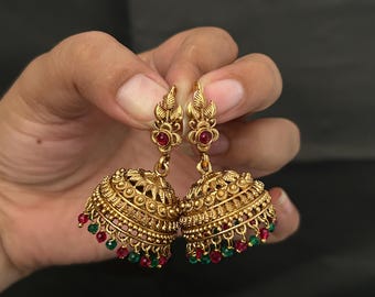 May include: A pair of gold-toned jhumka earrings featuring intricate metalwork, a central red stone on the stud, and a bell-shaped dome adorned with small red and green beads hanging from the bottom edge of the ornate metal structure.