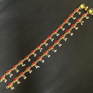 May include: A pair of gold-toned metal anklets featuring a repeating pattern of red enamel lotus flower charms. Each lotus charm is accented with small, dangling white beads. The anklets are finished with a simple hook clasp at the ends.