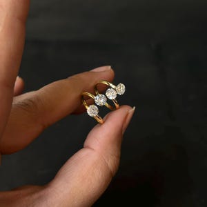 Op de afbeelding: Twee gouden ringen, elk met een grote, heldere, ronde edelsteen. De ringen worden tussen de vingers gehouden tegen een donkere achtergrond, wat hun heldere, reflecterende kwaliteit benadrukt. De gouden banden buigen elegant.