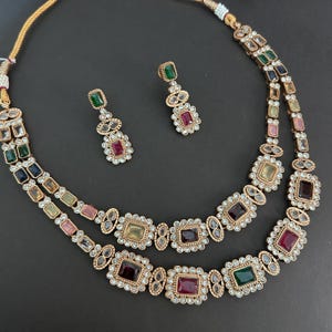 Puede incluir: Un conjunto de collar y pendientes de color dorado. El collar presenta dos hileras de piedras preciosas cuadradas y ovaladas de varios colores, incluyendo rojo, verde y amarillo. Los pendientes reflejan el diseño del collar.