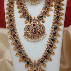 以下が含まれることがあります： ネックレス、イヤリング、ペンダントを含む、豪華な金色のジュエリーセット。 ネックレスとイヤリングには赤と緑の宝石が飾られ、ペンダントには孔雀のデザインがあります。 セットは白いスタンドに展示されています。