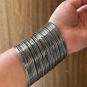 May include: A collection of silver-coloured bangles on a wrist. The bangles are thin, round, and have a shiny, metallic finish. The bracelets are stacked closely together, creating a bold and eye-catching look.