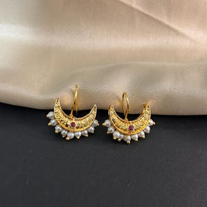 May include: A pair of gold-colored crescent moon earrings. Each earring features a small, round, red gemstone in the center and is adorned with small, white pearls along the bottom edge. The earrings have a textured design and a hook closure.