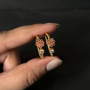 Könnte beinhalten: Ein Paar goldene Creolen mit einem strukturierten, seilartigen Band. Jeder Ohrring ist mit einer blumenförmigen Anordnung kleiner roter Steine und einem einzelnen klaren runden Stein unterhalb der Blume am Ring verziert.