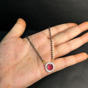 May include: A gold and black beaded necklace with a large, round, red gemstone pendant. The pendant is surrounded by a halo of small, clear stones. The necklace is displayed on a hand.