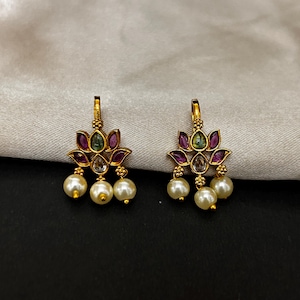 Puede incluir: Un par de pendientes dorados. Cada pendiente presenta un diseño floral con detalles morados y verdes, y tres perlas blancas colgantes. Los pendientes se muestran sobre un fondo neutro.