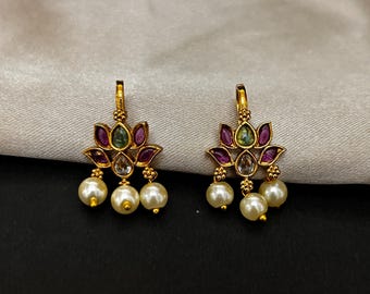 Pode incluir: Um par de brincos dourados. Cada brinco apresenta um design floral com detalhes roxos e verdes, e três pérolas brancas penduradas. Os brincos são exibidos em um fundo neutro.
