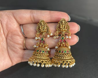 May include: A pair of gold-colored jhumka earrings. Each earring features a detailed deity figure at the top, with red, green, and white accents. The bell-shaped bottom is adorned with small white pearls, creating an elegant and traditional look.