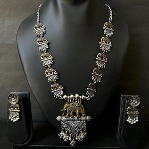 K&ouml;nnte beinhalten: Ein silberfarbenes Halsketten- und Ohrring-Set mit Elefantenmotiv. Die Halskette hat eine Kette mit sich wiederholenden Elefantenanh&auml;ngern und einen gr&ouml;&szlig;eren Anh&auml;nger mit zwei Elefanten. Die Ohrringe haben ein passendes Design. Der Schmuck wird auf einer schwarzen Schaufensterpuppe pr&auml;sentiert.