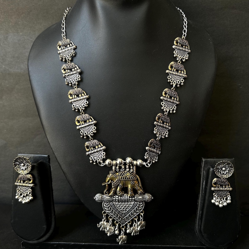 K&ouml;nnte beinhalten: Ein silberfarbenes Halsketten- und Ohrring-Set mit Elefantenmotiv. Die Halskette hat eine Kette mit sich wiederholenden Elefantenanh&auml;ngern und einen gr&ouml;&szlig;eren Anh&auml;nger mit zwei Elefanten. Die Ohrringe haben ein passendes Design. Der Schmuck wird auf einer schwarzen Schaufensterpuppe pr&auml;sentiert.