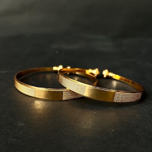 May include: Two gold bangles with a textured dotted pattern and a smooth rectangular centre. The bangles are displayed on a dark surface, showcasing their shiny, reflective quality. The jewellery is simple and elegant.