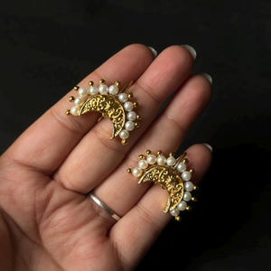 Op de afbeelding: Een paar goudkleurige, halvemaanvormige oorbellen met ingewikkelde filigraanontwerpen. Elke oorbel is versierd met kleine, witte, parelachtige kralen langs de gebogen rand, wat een delicate en elegante uitstraling creëert. De oorbellen worden tegen een donkere achtergrond gepresenteerd.