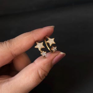 May include: Gold-coloured ring with three star designs. Two stars are embellished with small, clear gemstones, and the third is plain. The ring is displayed against a dark backdrop, accentuating its intricate details.