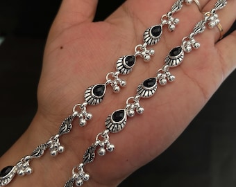 May include: A pair of silver-toned anklets featuring repeating teardrop-shaped black stones set in ornate metal frames. Each link is accented with small, dangling silver-toned spherical charms, creating a delicate and rhythmic design along the chain.