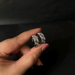 May include: Silver hoop earrings with a textured design. The earrings are embellished with small, sparkling stones. The design features a woven or braided pattern. The earrings are held against a dark background.
