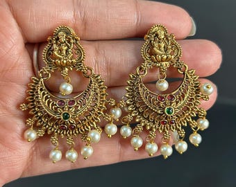 May include: Ornate gold-toned earrings featuring a crescent moon design with dangling faux pearls. Each earring has a small figure at the top, with red and green accents. The earrings are designed with intricate filigree details.