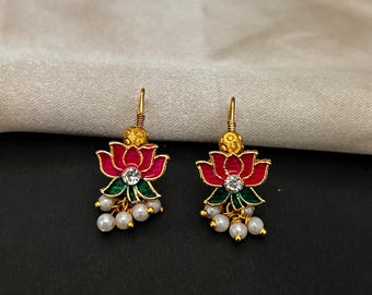 Pode incluir: Um par de brincos dourados com flores de lótus. Os brincos apresentam pétalas vermelhas com detalhes verdes, uma pedra central transparente e pérolas brancas penduradas. Os brincos têm um gancho dourado e uma pequena bola dourada no topo.