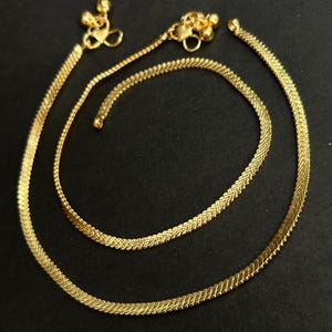 May include: A pair of gold-toned anklets featuring a flat, herringbone-style chain link design. Each anklet is finished with a small, decorative charm cluster consisting of tiny metallic spheres and a heart-shaped element at the clasp end.
