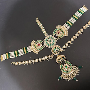 May include: An elaborate gold-coloured headpiece featuring green and red gemstones, white pearls, and a dangling pendant. The design incorporates floral and geometric patterns, with beaded and solid sections. The jewellery is set against a dark background.