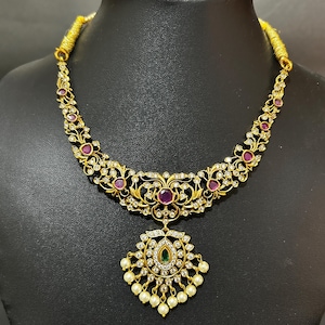 Puede incluir: Un collar dorado con un diseño floral, con piedras preciosas rojas y transparentes. El colgante tiene forma de lágrima con una gema verde y perlas blancas. El collar se exhibe en un maniquí negro.