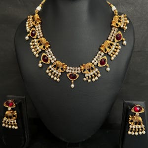 Può includere: Un set composto da collana e orecchini color oro con motivi a forma di elefante, pietre sfaccettate rosso scuro e piccole perline bianche. La collana presenta un ciondolo centrale con pietra rossa e perline pendenti coordinate.