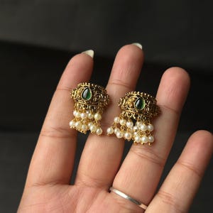 Op de afbeelding: Een paar goudkleurige oorbellen met een ingewikkeld metaalontwerp en een centrale druppelvormige groene steen. Elke oorbel is versierd met een trosje kleine, ronde witte parels die in een gelaagde opstelling aan de onderkant hangen.