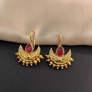May include: A pair of gold-colored earrings with a crescent moon design. Each earring features a teardrop-shaped red gemstone and intricate filigree detailing. Small gold beads adorn the bottom of the crescent.