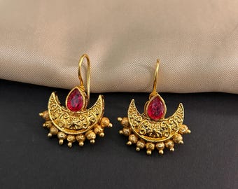 Pode incluir: Um par de brincos dourados com um design de meia-lua. Cada brinco apresenta uma pedra preciosa vermelha em forma de gota e detalhes intrincados em filigrana. Pequenas contas douradas adornam a parte inferior da meia-lua.