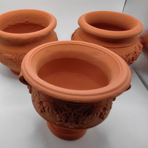 Può includere: Tre vasi in terracotta di colore marrone-rossastro. I vasi hanno un bordo ampio e arrotondato e un motivo floreale decorativo attorno alla base. I vasi sono adatti per piantare fiori o erbe.