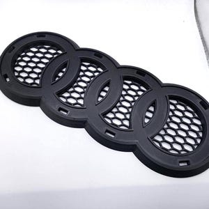 May include: A black Audi logo, comprising four interlocking rings with a honeycomb mesh design. The logo is three-dimensional and made of a matte black material. It is set against a white background. The design is a car emblem.