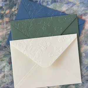 May include: Three envelopes in shades of blue, green, and cream, arranged in a layered composition. The cream envelope features a floral embossed design, while the other two have a subtle floral pattern. The envelopes are likely for invitations or stationery.