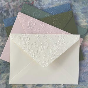May include: A collection of floral-embossed envelopes in various pastel colors, including pink, green, and blue. The envelopes are arranged in a fan shape, showcasing the delicate floral pattern on the flaps. The background is a textured, multi-colored surface.
