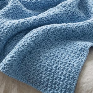 May include: A light blue blanket featuring a dense, textured crochet stitch pattern. The fabric displays a consistent, repeating knot-like weave throughout the entire surface, creating a thick, soft appearance with visible depth and dimension.