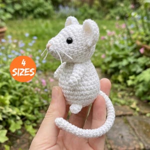 Könnte beinhalten: Eine kleine, weiß gehäkelte Mausfigur mit schwarzen Perlenaugen, einer rosa gestickten Nase und langen weißen Schnurrhaaren. Die Maus hat abgerundete Ohren, einen runden Körper und einen langen, geschwungenen Schwanz. Ein orangefarbener Kreis zeigt 4 SIZES.
