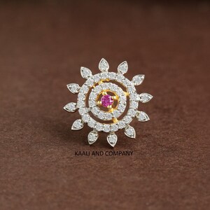 May include: A circular gold nose stud featuring a central round pink gemstone surrounded by two concentric rings of small white stones and an outer border of petal-shaped white stones. The text KAAU AND COMPANY is printed below the stud.