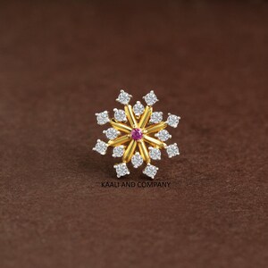 May include: A gold nose stud shaped like a snowflake, featuring a central small pink gemstone surrounded by radiating gold spokes tipped with clear white stones. The text KAALI AND COMPANY is printed in small black letters below the jewelry piece.