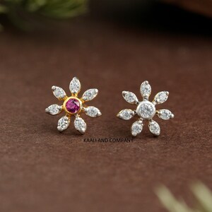 May include: A pair of flower-shaped stud earrings. One earring features a central red gemstone, while the other has a clear gemstone. Both earrings have white, petal-shaped accents and gold-colored settings. The earrings are displayed on a brown surface.