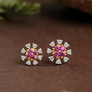 May include: A pair of gold-toned stud earrings. Each earring features a central cluster of pink gemstones surrounded by white gemstones, creating a floral design. The earrings are set against a brown background.