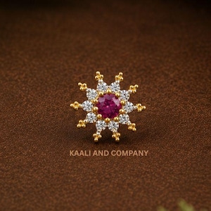 Könnte beinhalten: Ein goldfarbener Nasenpiercing mit einem zentralen Rubin, umgeben von weißen und goldenen Akzenten. Der Piercing hat ein Sternmuster, darunter der Text "KAALI AND COMPANY". Der Hintergrund ist braun mit grünem Laub.