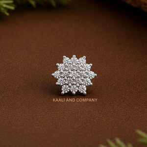 May include: A cluster of small, round, clear gemstones arranged in a starburst pattern. The jewelry piece is set against a brown background with the text "KAALI AND COMPANY" at the bottom.