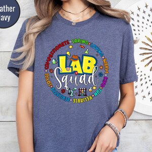 Pode incluir: Camiseta azul marinho mesclada com um gráfico colorido "LAB Squad". O design apresenta um círculo amarelo e azul com palavras como "compassivo" e "confiável". A camiseta tem mangas curtas e gola redonda.