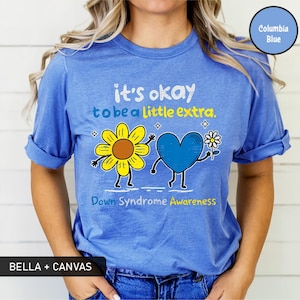 May include: Columbia blue t-shirt with the text "it's okay to be a little extra." and "Down Syndrome Awareness." The graphic features a yellow sunflower and a blue heart holding a daisy. The shirt is made by Bella + Canvas.