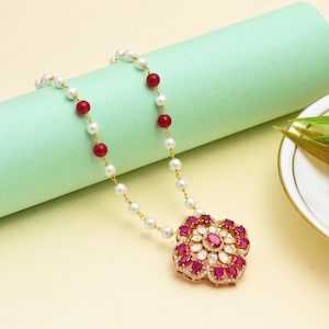Könnte beinhalten: Eine goldene Halskette mit einem Anhänger in Blumenform, verziert mit roten Edelsteinen und weißen Akzenten. Die Kette besteht aus weißen Perlen, roten Perlen und goldenen Gliedern. Der Schmuck wird vor einem hellgelben und grünen Hintergrund präsentiert.