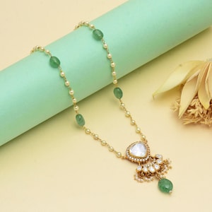 Op de afbeelding: Goudkleurige ketting met een hanger met een grote, heldere steen en kleinere witte stenen. De ketting heeft een delicate ketting afgewisseld met kleine parels en groene edelsteen kralen. De hanger is geaccentueerd met kleine parels en een groene edelsteen druppel.