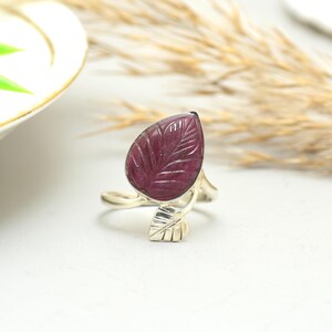 Könnte beinhalten: Ein Silberring mit einem geschnitzten, blattförmigen, tiefroten Edelstein. Das Design des Rings umfasst ein detailliertes Blattmotiv am Band, wobei der Edelstein etwa 2.5 cm lang ist.