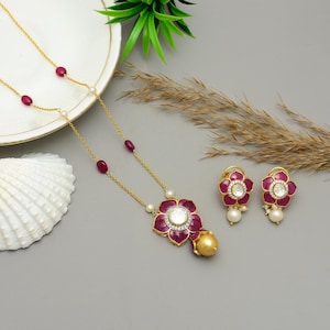 Könnte beinhalten: Ein goldfarbenes Halsketten- und Ohrring-Set. Die Halskette hat einen Blumenanhänger mit einem zentralen weißen Stein, umgeben von einem roten Emaille-Blumenmuster. Die Ohrringe spiegeln das Anhängerdesign wider. Das Set enthält rote und weiße Edelsteine.