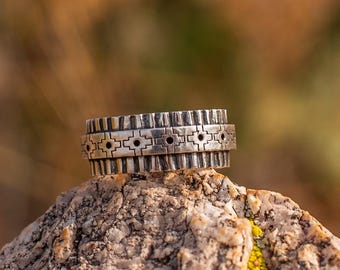 May include: A silver ring with a textured design. The ring features a series of rectangular and geometric patterns. The ring is displayed on a rock with a blurred background.
