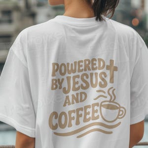 Puede incluir: Camiseta blanca con el texto "POWERED BY JESUS AND COFFEE" en fuente marrón retro. El diseño incluye una cruz y una taza de café humeante. La camiseta tiene mangas cortas.