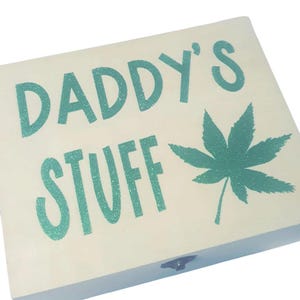 May include: A rectangular wooden box with the words "DADDY'S STUFF" in teal glitter lettering. A teal glitter cannabis leaf is on the right side of the box. The box is off-white.